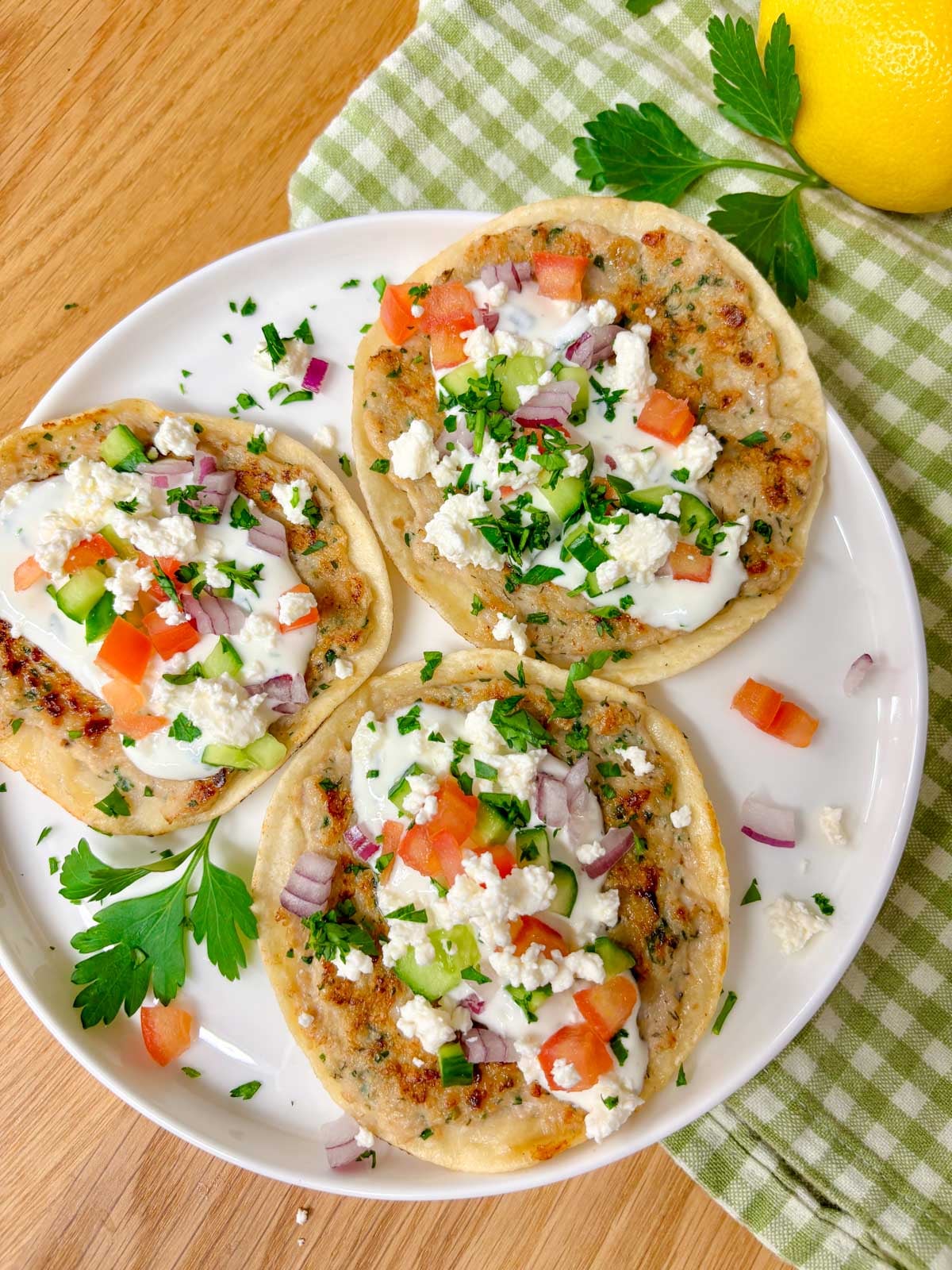 a plate is filled with three greek chicken smash tacos topped with tzatziki, onion, tomato, cucumber, onion, feta, and parsley