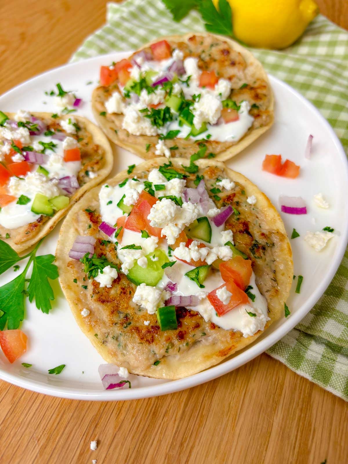a plate is filled with three greek chicken smash tacos topped with tzatziki, onion, tomato, cucumber, onion, feta, and parsley