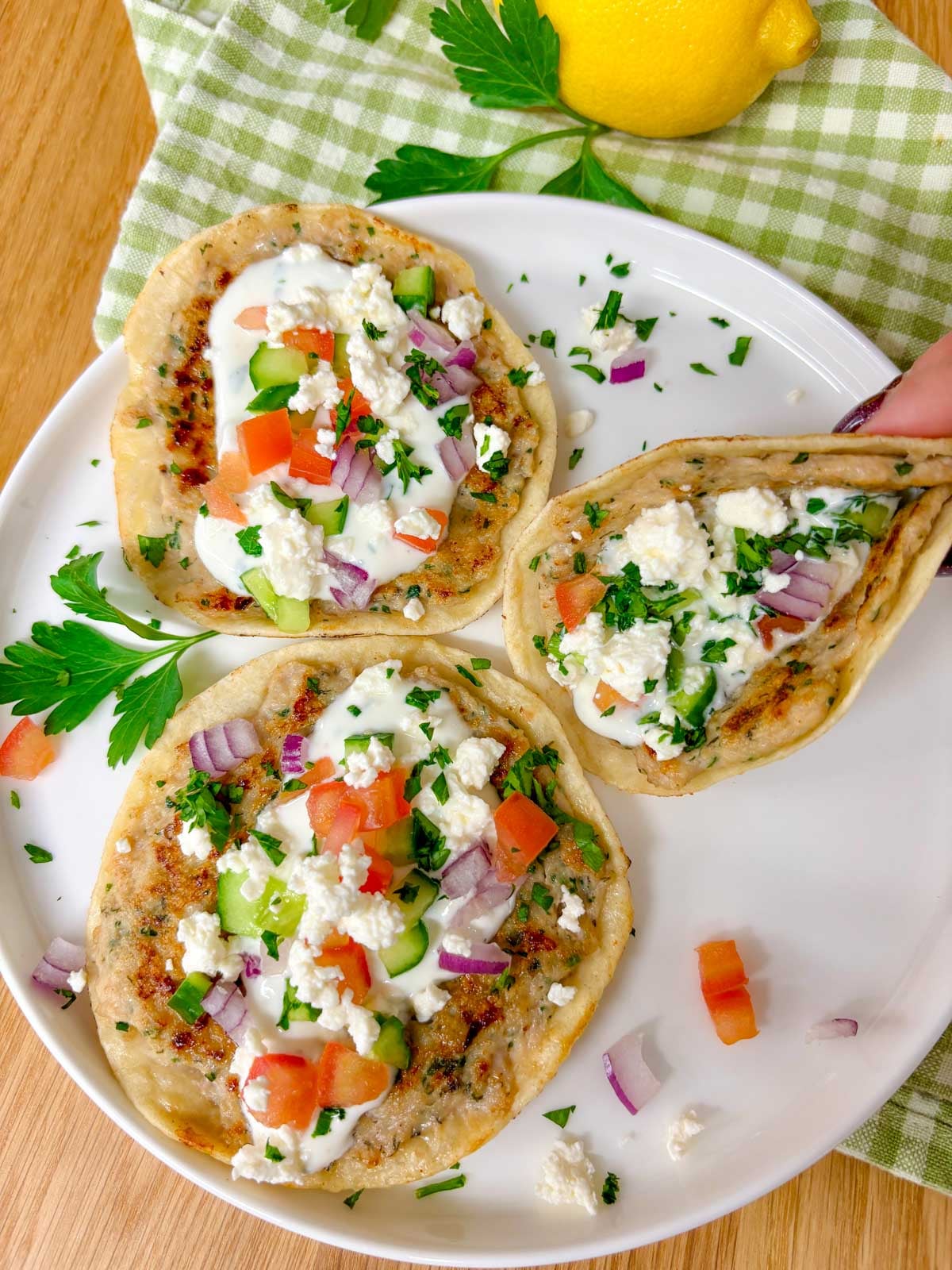 a plate is filled with three greek chicken smash tacos topped with tzatziki, onion, tomato, cucumber, onion, feta, and parsley. one is folded in half