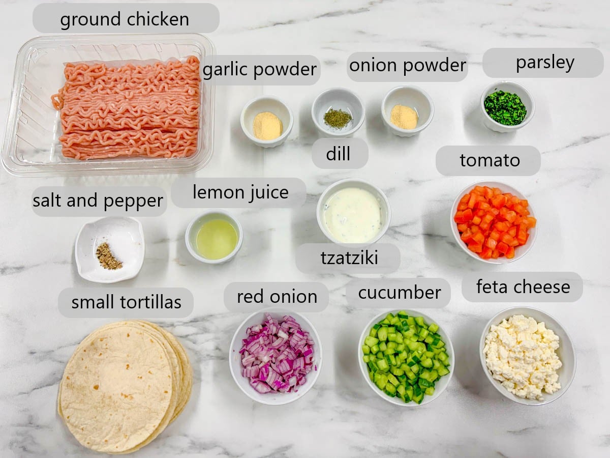kitchen counter with ingredients in dishes and text overlays reading ground chicken, garlic powder, dill, onion powder, parsley, salt and pepper, lemon juice, tzatziki, tomato, small tortillas, red onion, cucumber, and feta cheese.