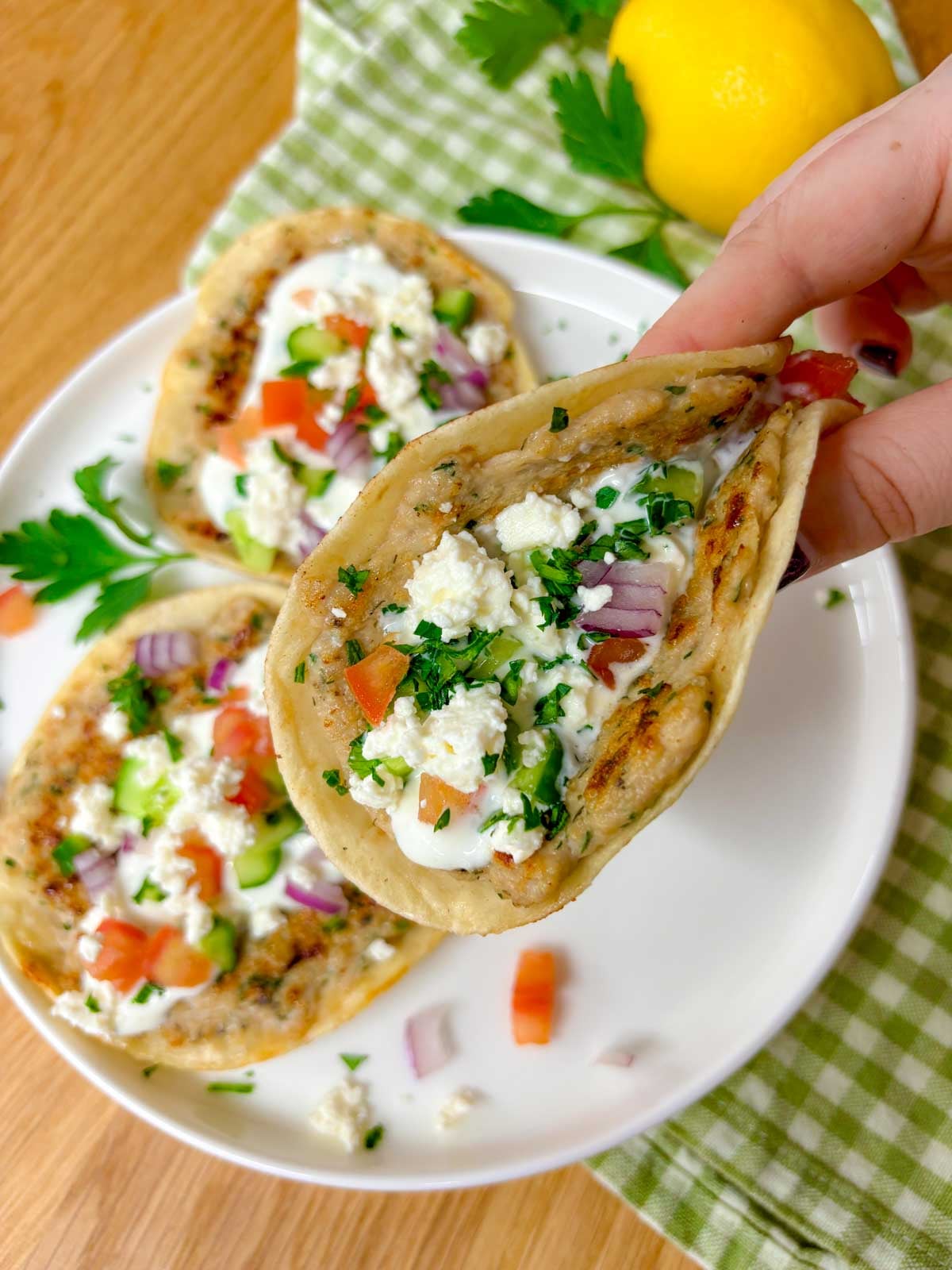 a plate is filled with three greek chicken smash tacos topped with tzatziki, onion, tomato, cucumber, onion, feta, and parsley. one is held and folded in half