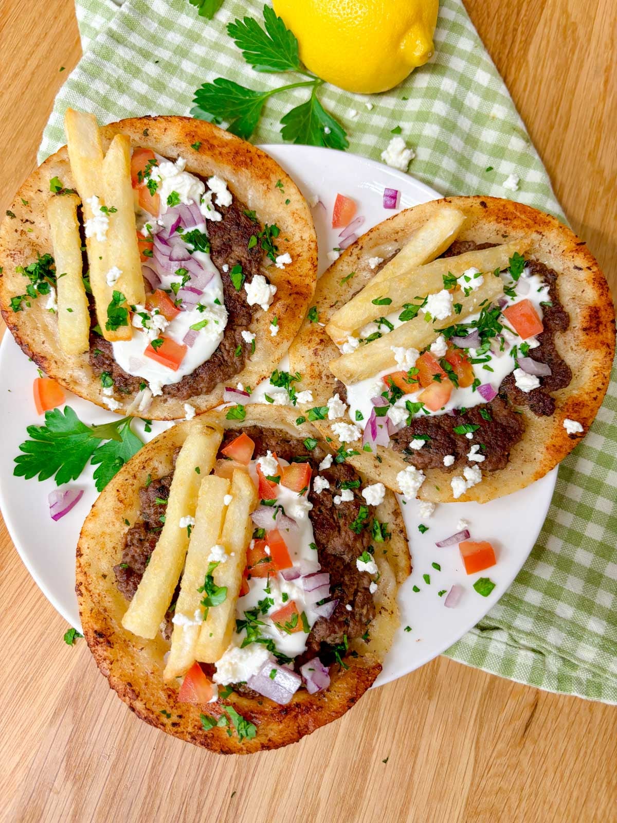 a white plate is filled with three Greek gyros smash tacos with tzatziki, fries, tomato, onion, and feta on top
