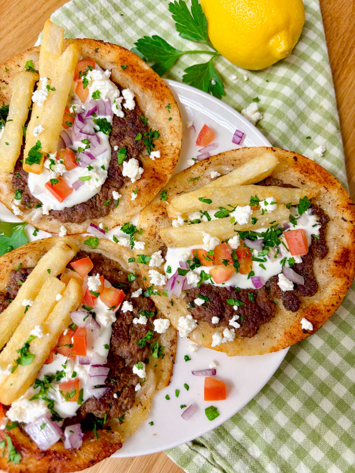 a white plate is filled with three Greek gyros smash tacos with tzatziki, fries, tomato, onion, and feta on top