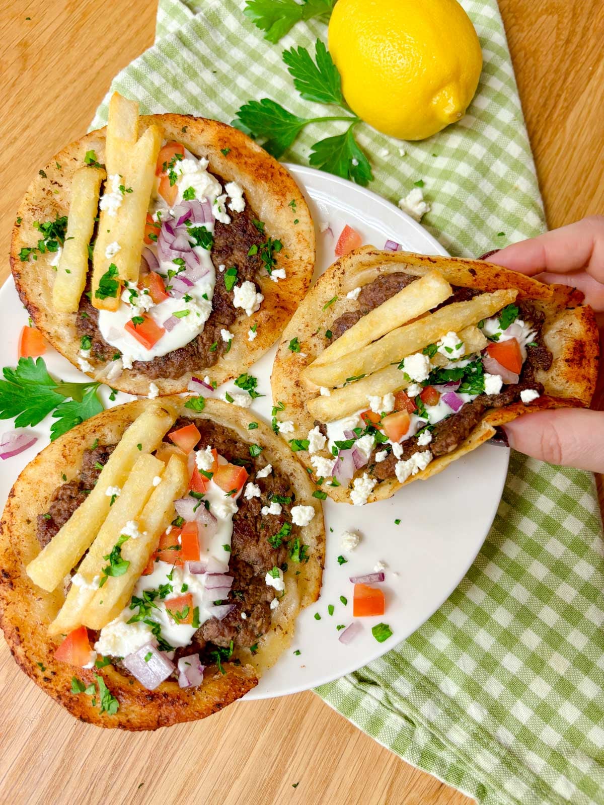a white plate is filled with three Greek gyros smash tacos with tzatziki, fries, tomato, onion, and feta on top. one is held