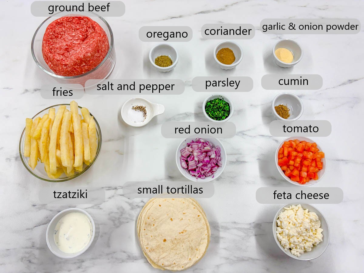 kitchen counter with ingredients and text overlays reading ground beef, oregano, coriander, garlic & onion powder, fries, salt and pepper, parsley, cumin, red onion, tomato, tzatziki, small tortillas, feta cheese
