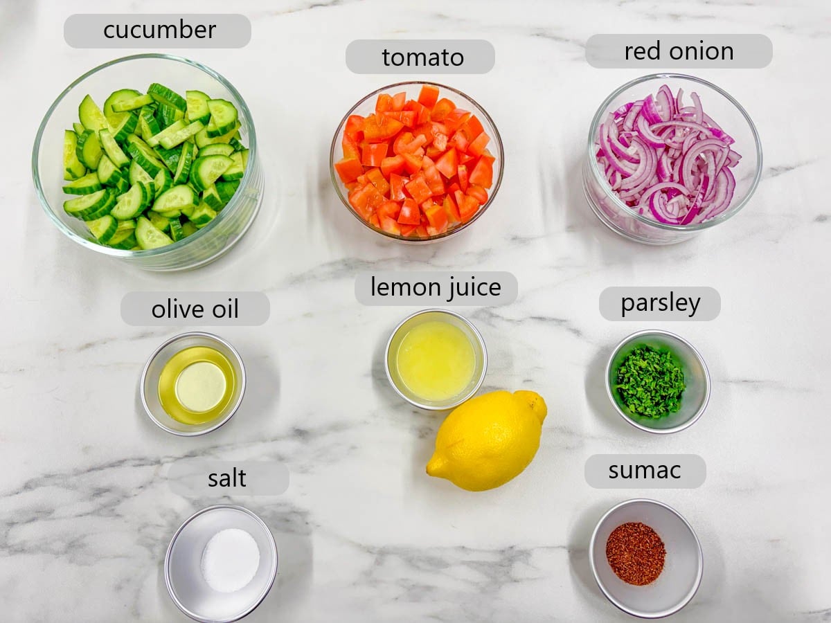 kitchen counter with bowls of ingredients with text overlays reading cucumber, tomato, red onion, olive oil, lemon juice, parsley, salt, and sumac.