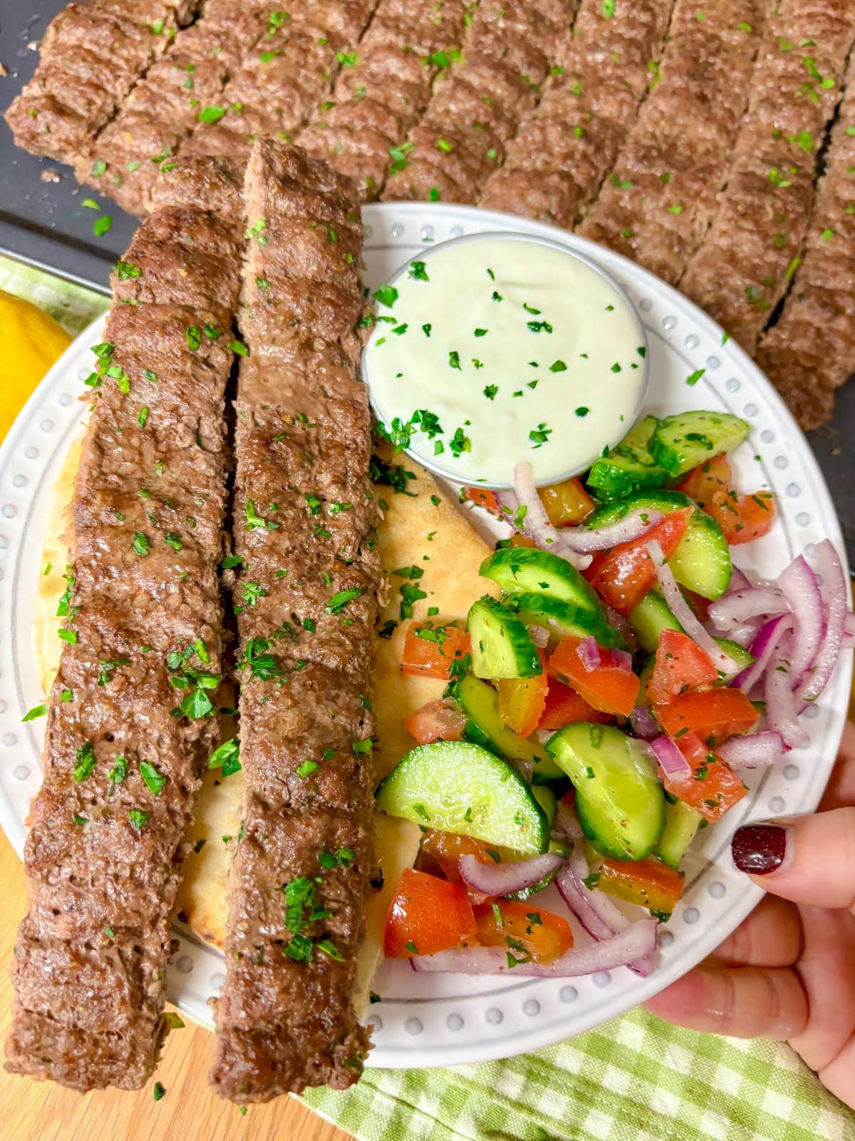 holding a plate of beef kebab dinner and in the background the sheet pan of keba bs