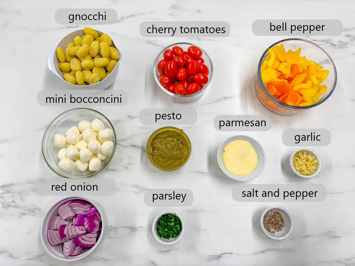 a kitchen counter with bowls of ingredients and text overlays reading gnocchi, cherry tomatoes, bell peppers, mini bocconcini, pesto, parmesan, garlic, red onion, parsley, salt and pepper