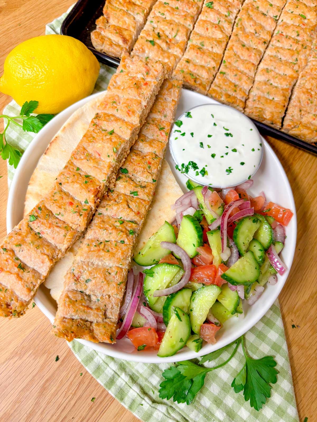 a plate is topped with pita bread, two chicken kebabs, cucumber salad, and a ramekin of garlic sauce.