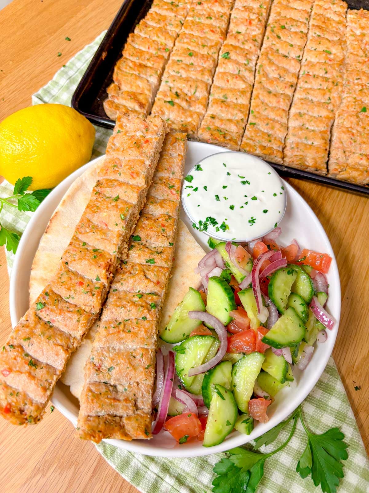 a plate is topped with pita bread, two chicken kebabs, cucumber salad, and a ramekin of garlic sauce.