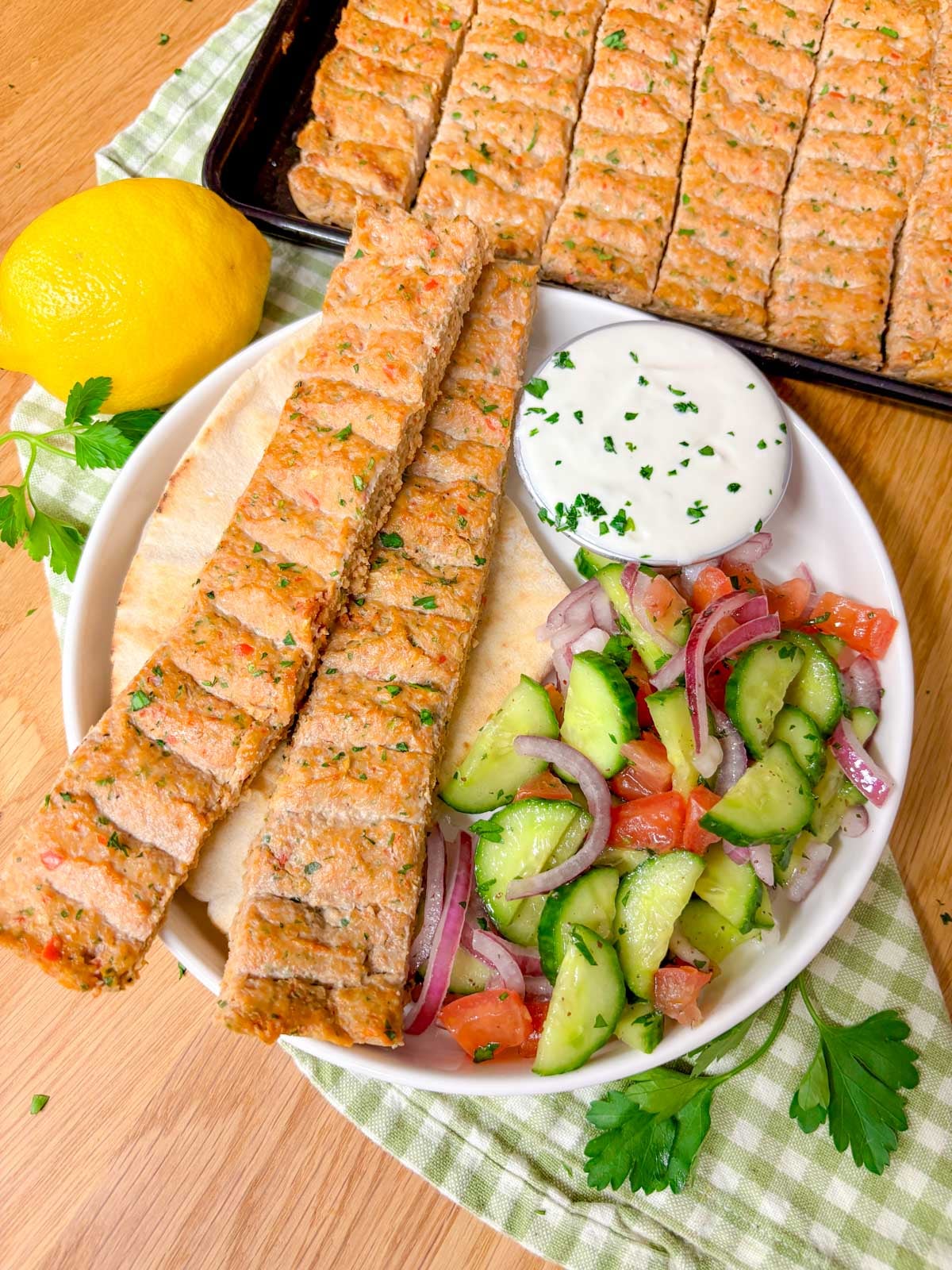 a plate is topped with the sheet pan chicken kebab dinner featuring pita bread, two chicken kebabs, cucumber salad, and a ramekin of garlic sauce.