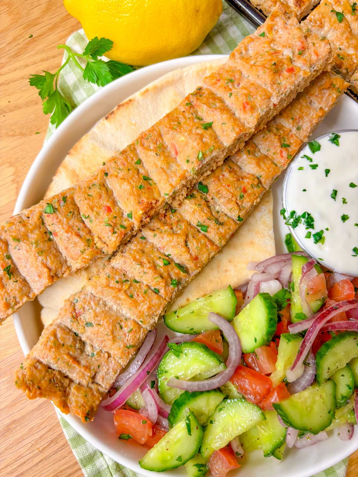 a plate is topped with pita bread, two chicken kebabs, cucumber salad, and a ramekin of garlic sauce.