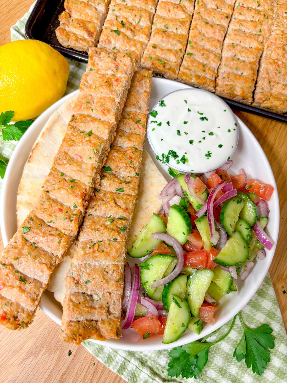 a plate is topped with pita bread, two chicken kebabs, cucumber salad, and a ramekin of garlic sauce.