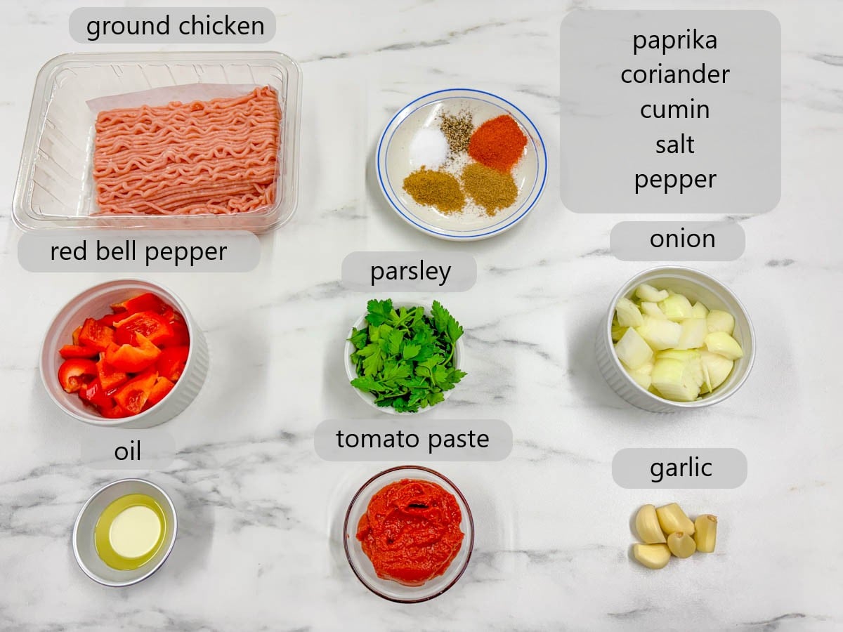 kitchen counter with bowls and ingredients with text overlays reading ground chicken, paprika, coriander, cumin, salt, pepper, red bell pepper, parsley, onion, oil, tomato paste, garlic.