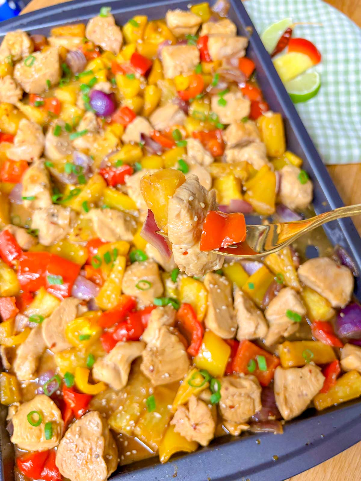 a sheet pan is filled with pineapple chicken stir fry and garnished with green onions and sesame seeds. a bite is on a fork