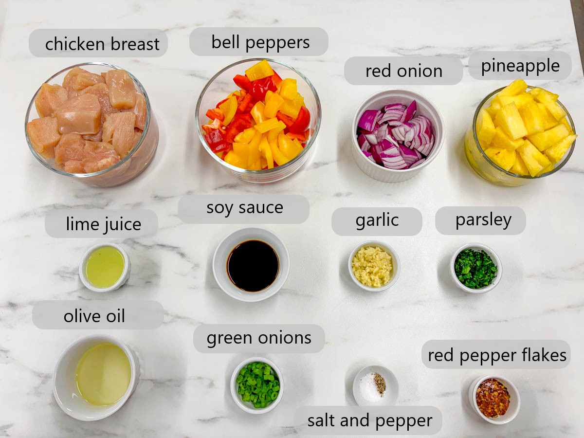 kitchen counter with bowls of ingredient and text overlays reading chicken breast, bell peppers, red onion, pineapple, lime juice, soy sauce, garlic, parsley, olive oil, green onions, salt and pepper, and red pepper flakes
