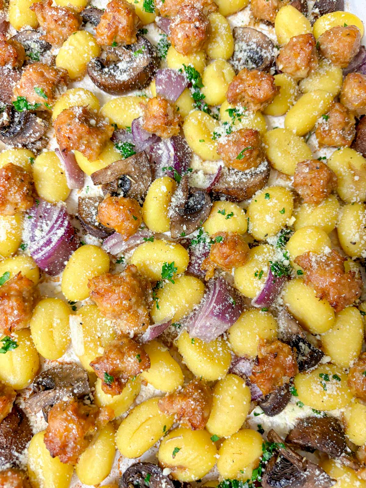 a sheet pan is filled with cooked gnocchi, sausage, mushrooms and red onion. It is garnished with parmesan cheese and parsley.