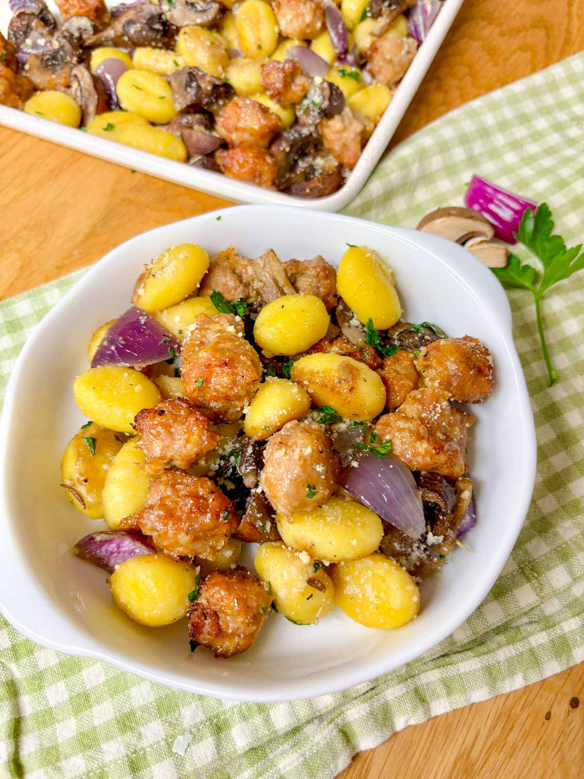 a sheet pan is filled with cooked gnocchi, sausage, mushrooms and red onion. It is garnished with parmesan cheese and parsley. a small bowl in front too.