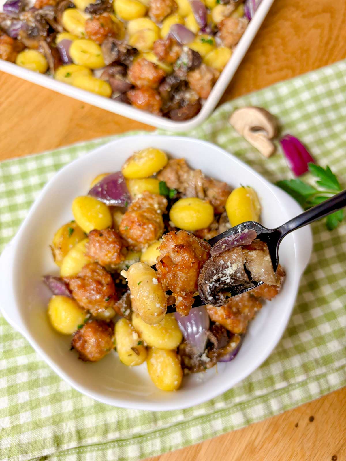 a sheet pan is filled with cooked gnocchi, sausage, mushrooms and red onion. It is garnished with parmesan cheese and parsley. a small bowl in front with a fork holding some food.