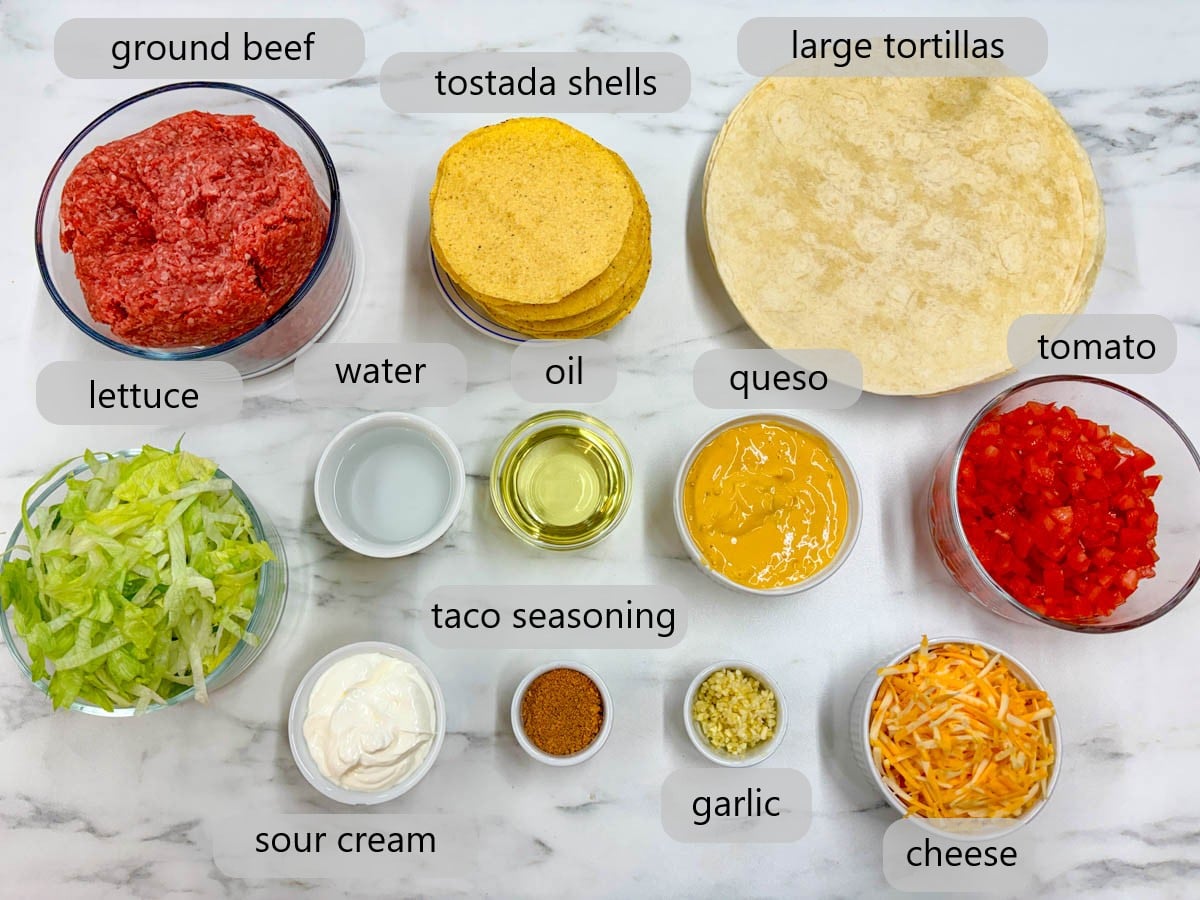 kitchen counter with bowls of ingredients and text overlays reading ground beef, tostada shells, large tortillas, lettuce, water, oil, queso, tomato, sour cream, taco seasoning, garlic, and cheese