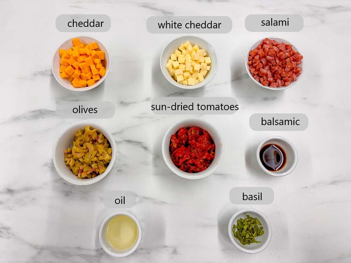 kitchen counter with bowls of ingredients and text overlays reading cheddar, white cheddar, salami, olives, sun-dried tomatoes, balsamic, oil, and basil.