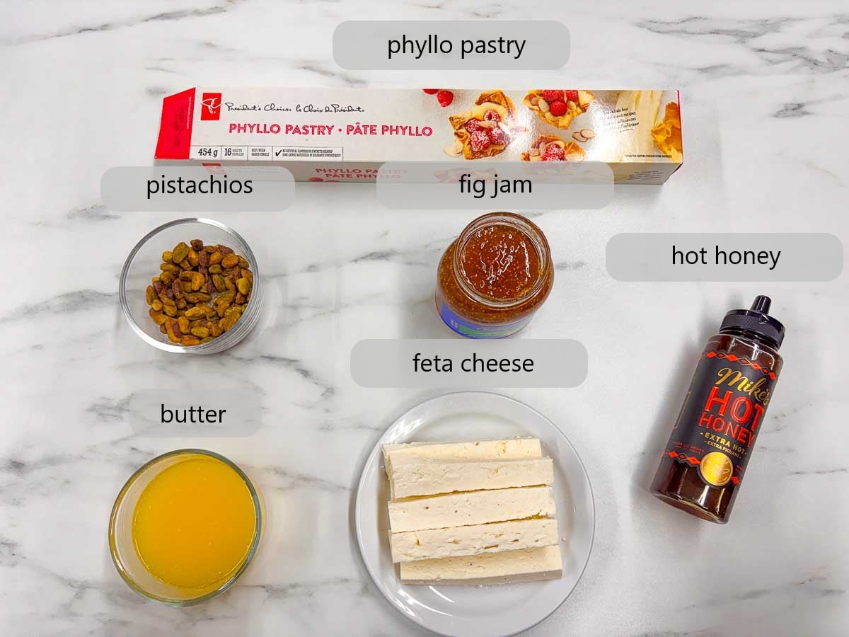 kitchen counter with ingredients and text overlays reading phyllo pastry, pistachios, fig jam, hot honey, butter, and feta cheese