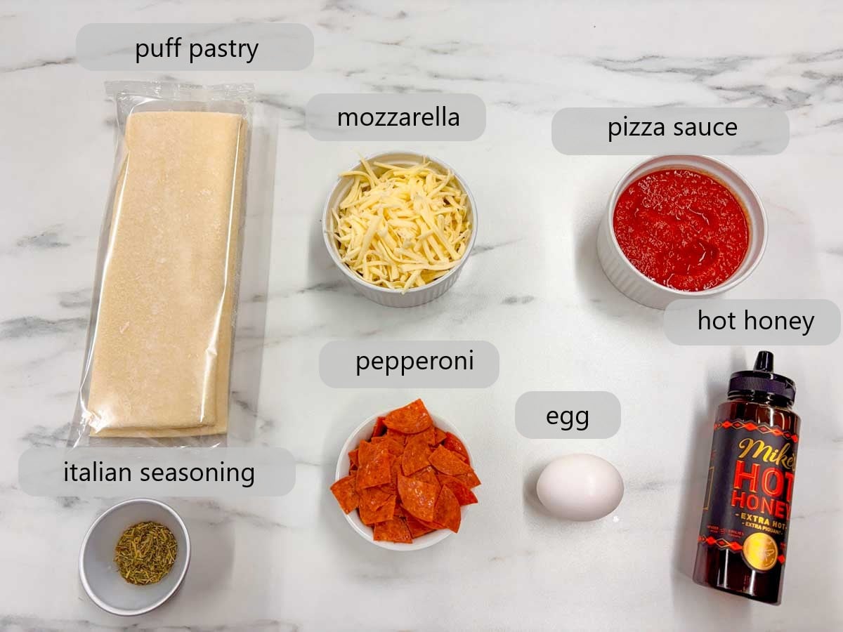 kitchen counter with ingredient and text overlays reading puff pastry, mozzarella, pizza sauce, Italian seasoning, pepperoni, egg, and hot honey.