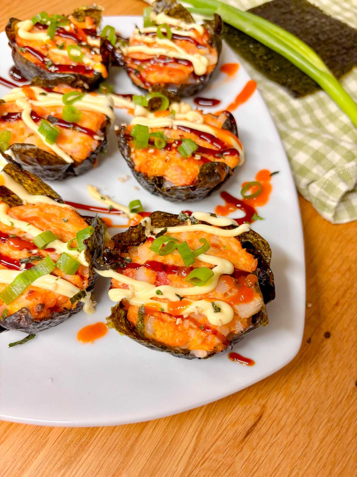 a white platter is filled with sushi bake cups and garnished with sriracha, kewpie mayo, eel sauce, green onions and furikake.