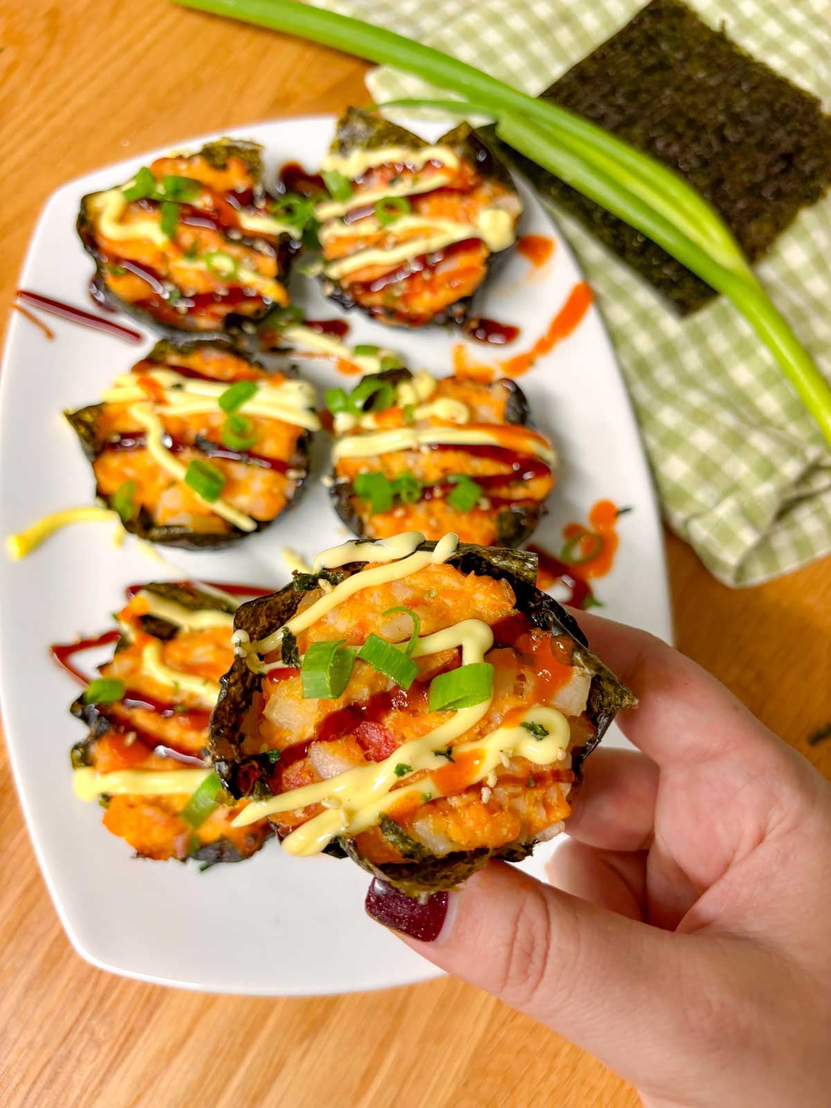 a white platter is filled with sushi bake cups and garnished with sriracha, kewpie mayo, eel sauce, green onions and furikake. one is help up close to camera