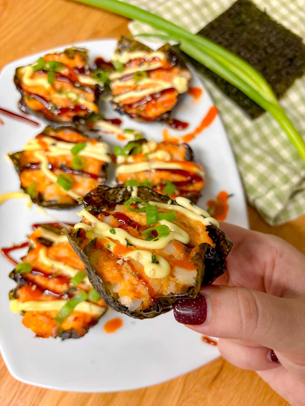 a white platter is filled with sushi bake cups and garnished with sriracha, kewpie mayo, eel sauce, green onions and furikake. one is held close to camera