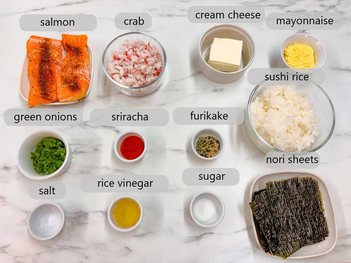 kitchen counter with bowls of ingredients and text overlays reading salmon, crab, cream cheese, mayonnaise, green onions, sriracha, furikake, sushi rice, salt, rice vinegar, sugar, and nori sheets.