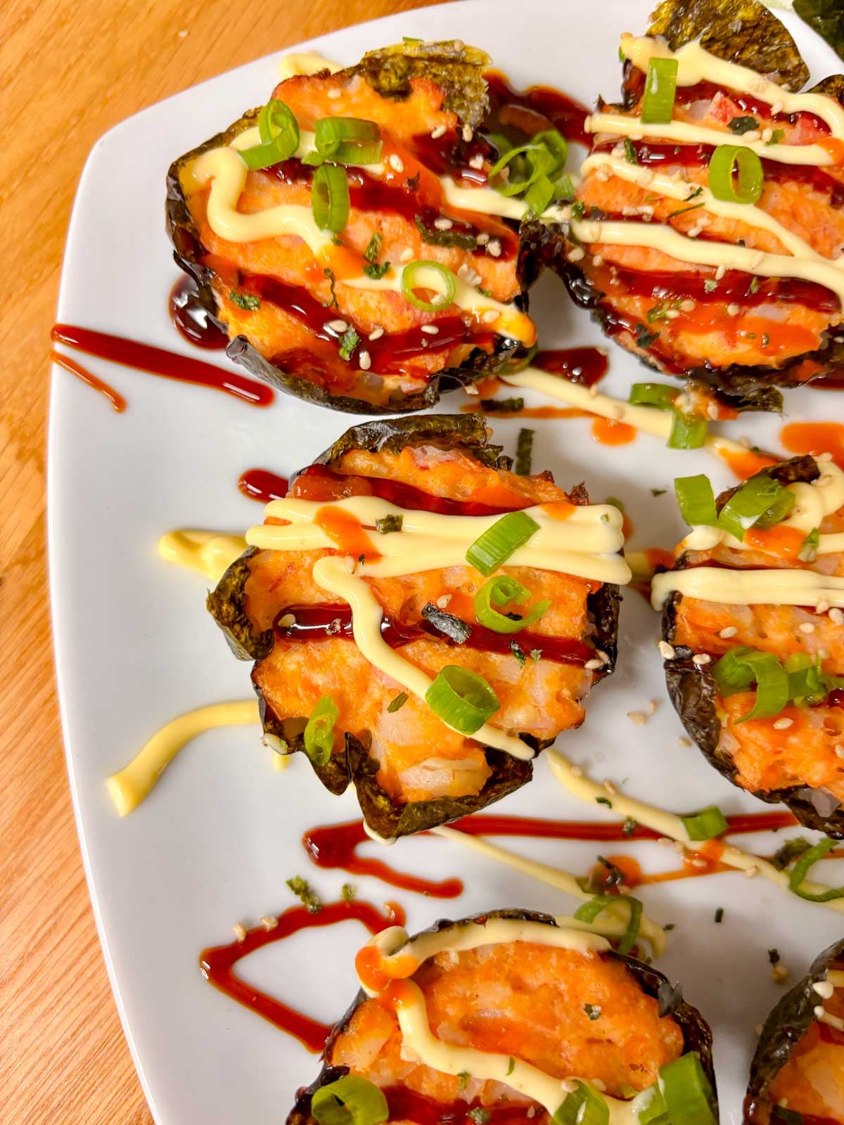 a white platter is filled with sushi bake cups and garnished with sriracha, kewpie mayo, eel sauce, green onions and furikake.