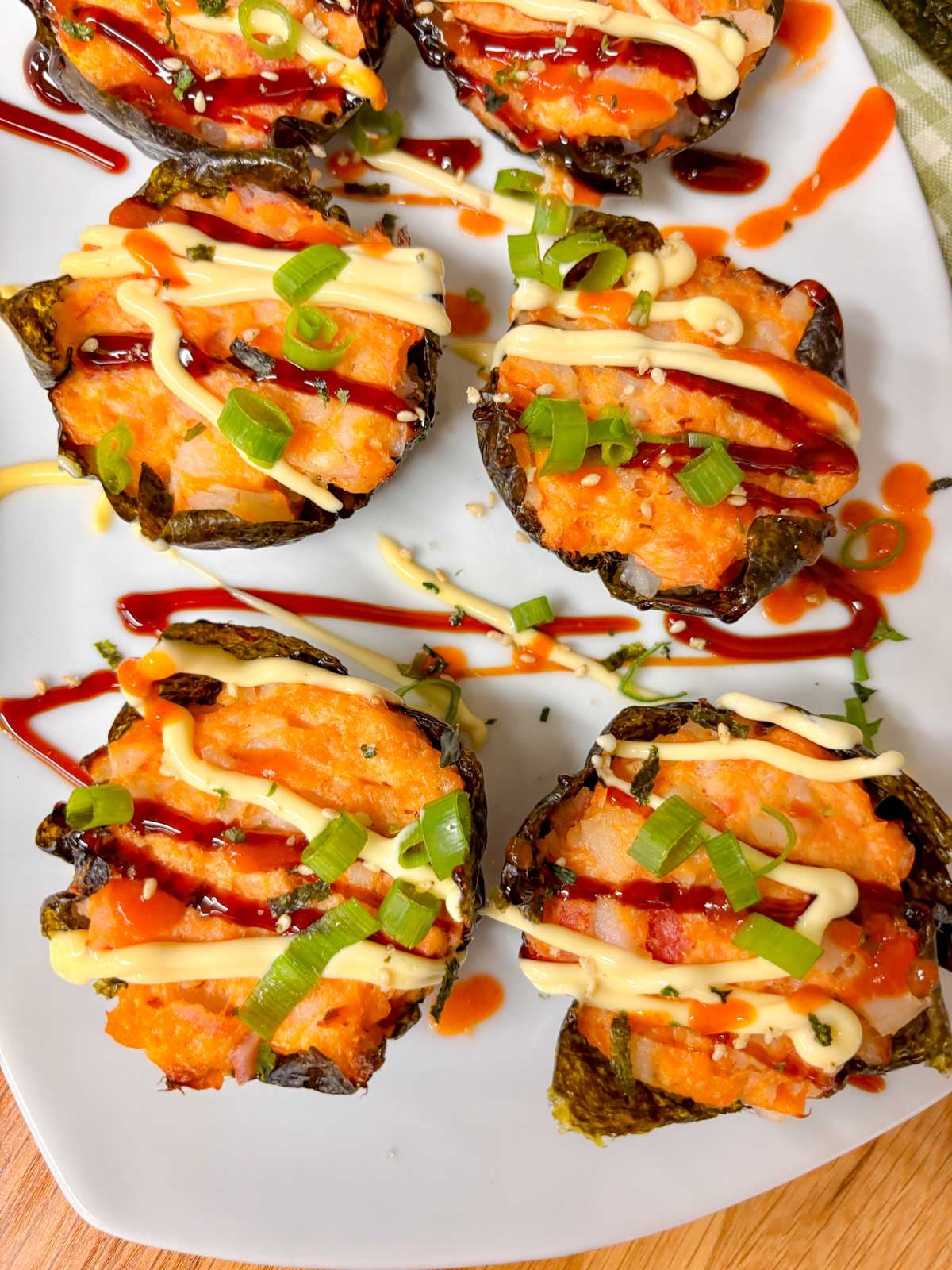 a white platter is filled with sushi bake cups and garnished with sriracha, kewpie mayo, eel sauce, green onions and furikake.