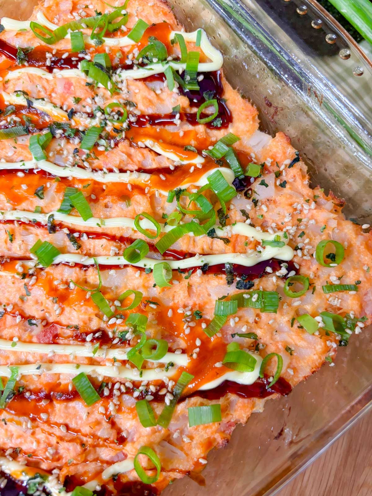 a square glass baking dish is filled with a spicy salmon and crab sushi bake. it is garnished with eel sauce, sriracha, kewpie mayo, green onions, and furikake.
