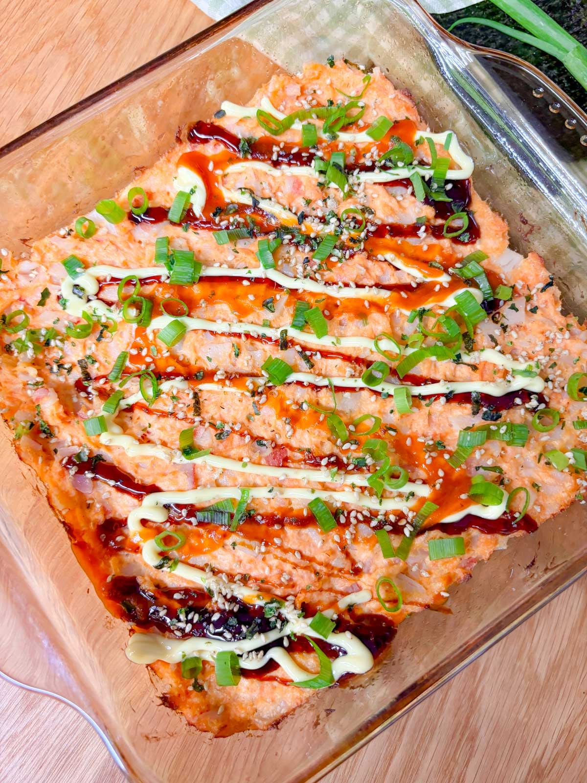a square glass baking dish is filled with a spicy salmon and crab sushi bake. it is garnished with eel sauce, sriracha, kewpie mayo, green onions, and furikake.