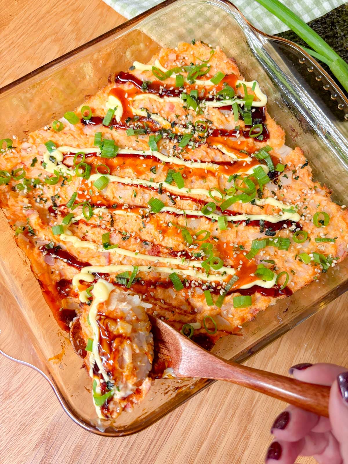 a square glass baking dish is filled with a spicy salmon and crab sushi bake. it is garnished with eel sauce, sriracha, kewpie mayo, green onions, and furikake. a spoon is scooping some of the sushi bake up.