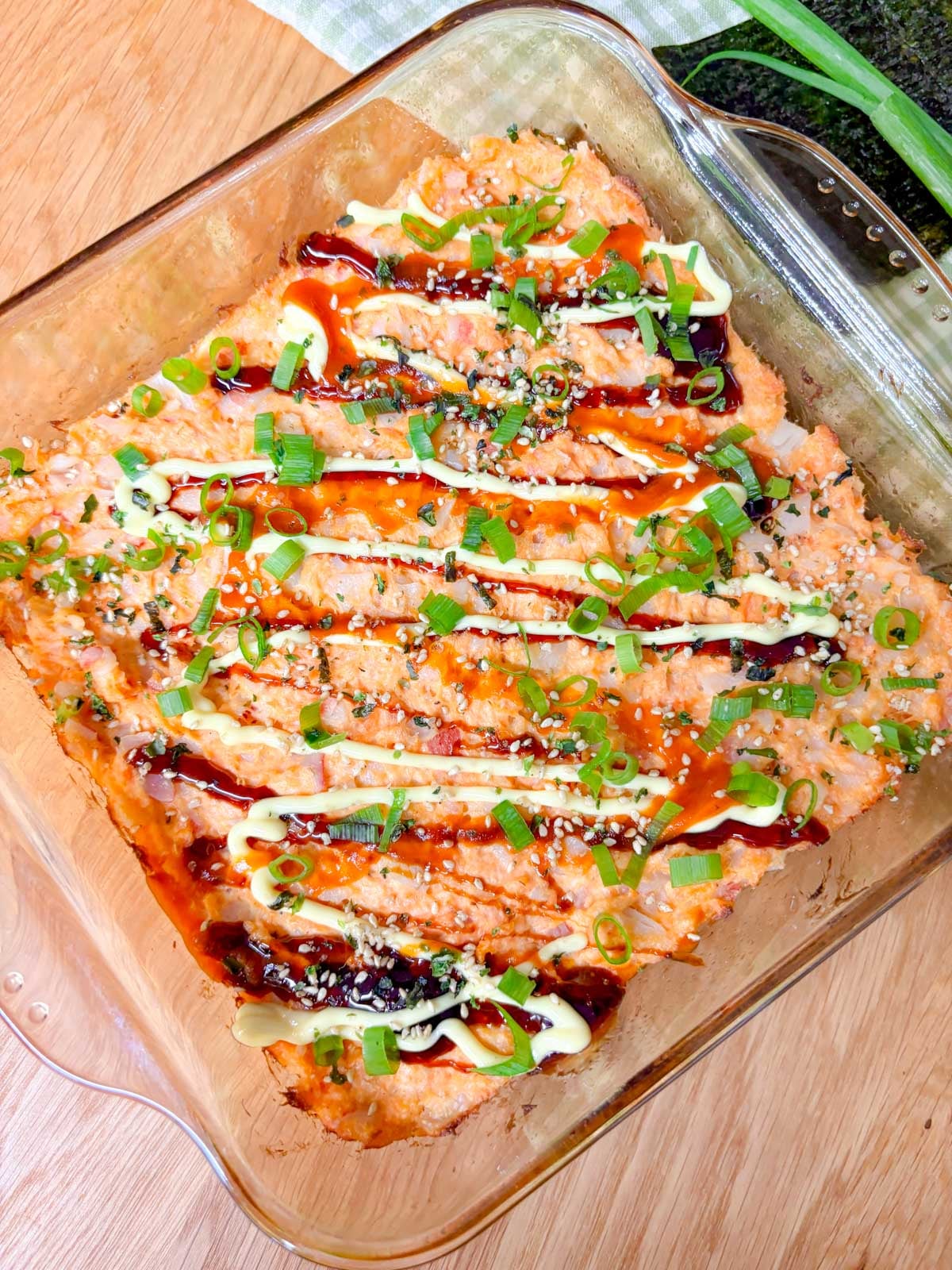 a square glass baking dish is filled with a spicy salmon and crab sushi bake. it is garnished with eel sauce, sriracha, kewpie mayo, green onions, and furikake.