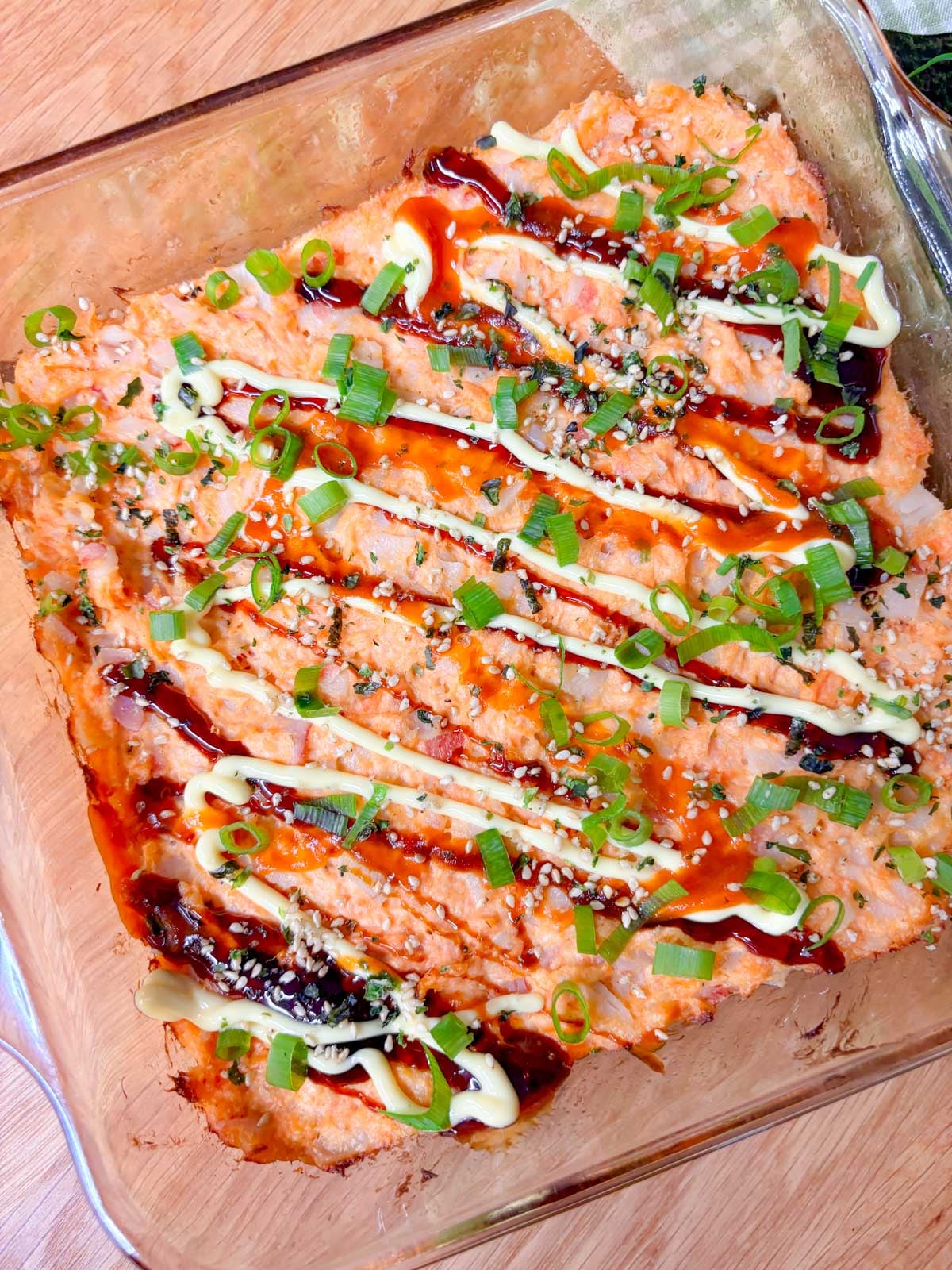 a square glass baking dish is filled with a spicy salmon and crab sushi bake. it is garnished with eel sauce, sriracha, kewpie mayo, green onions, and furikake.