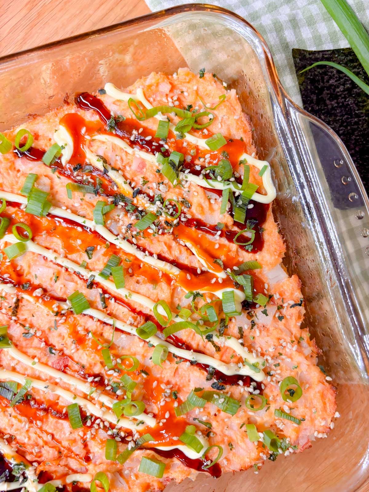 a square glass baking dish is filled with a spicy salmon and crab sushi bake. it is garnished with eel sauce, sriracha, kewpie mayo, green onions, and furikake.